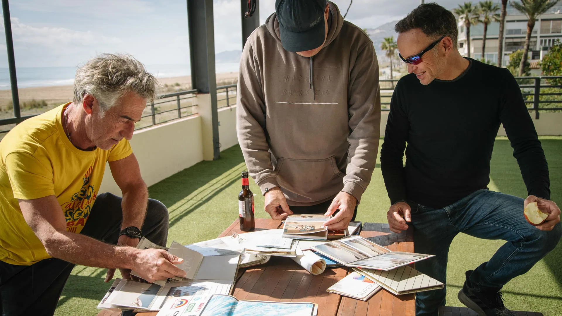265 días sin olas: tres surfistas repasan fotos y revistas antiguas en una terraza junto al mar