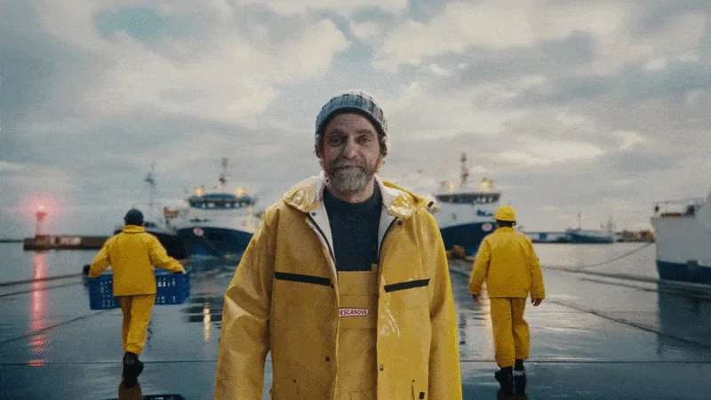 Pescanova commercial still: fisherman in yellow oilskins on a rainy dock, trawlers behind